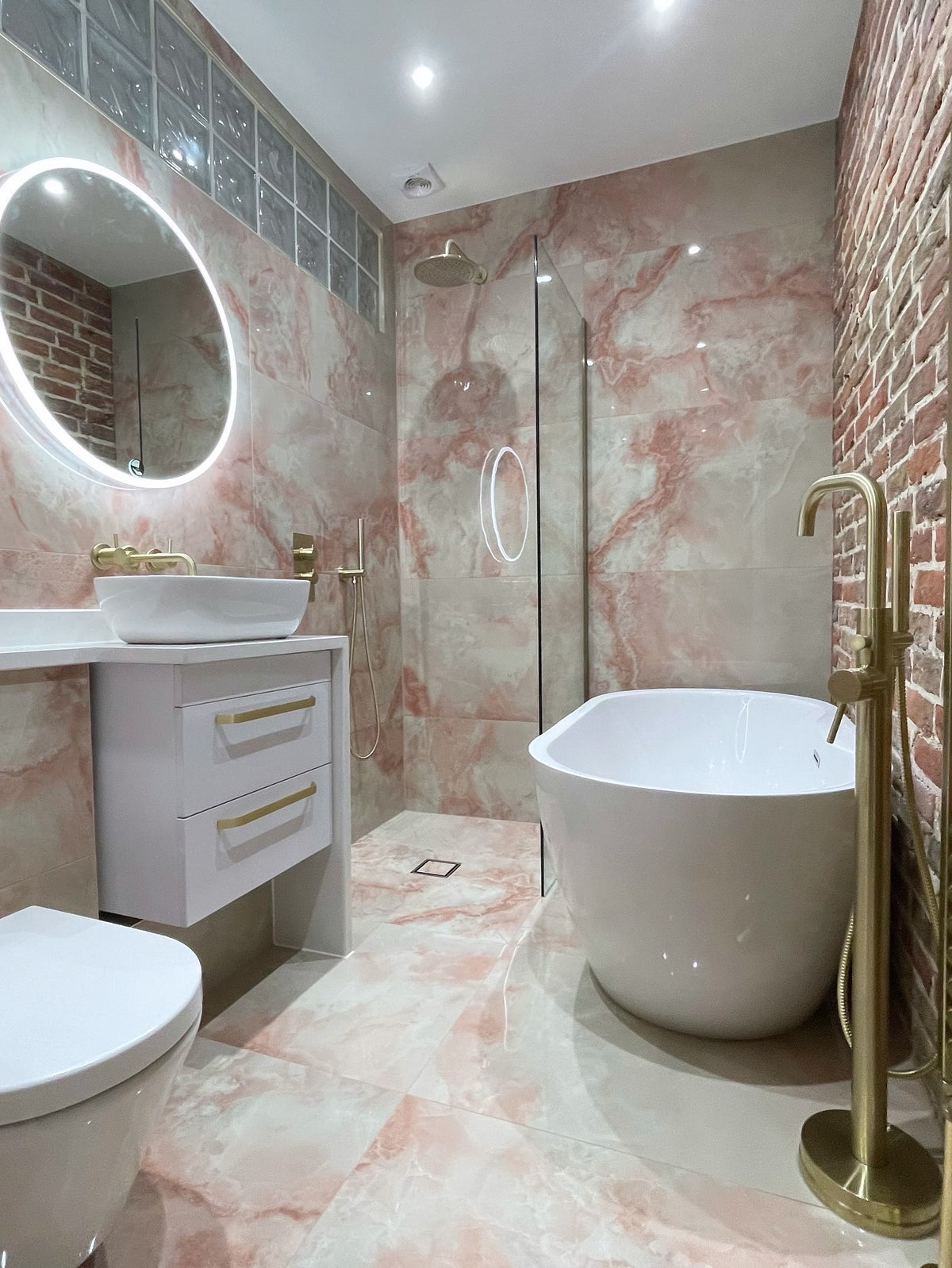 A modern bathroom interior featuring a freestanding white bathtub, a white vanity with a vessel sink and illuminated round mirror, a glass shower enclosure, pink marble-look tile walls and floor, exposed brick accents, and gold-tone fixtures.
