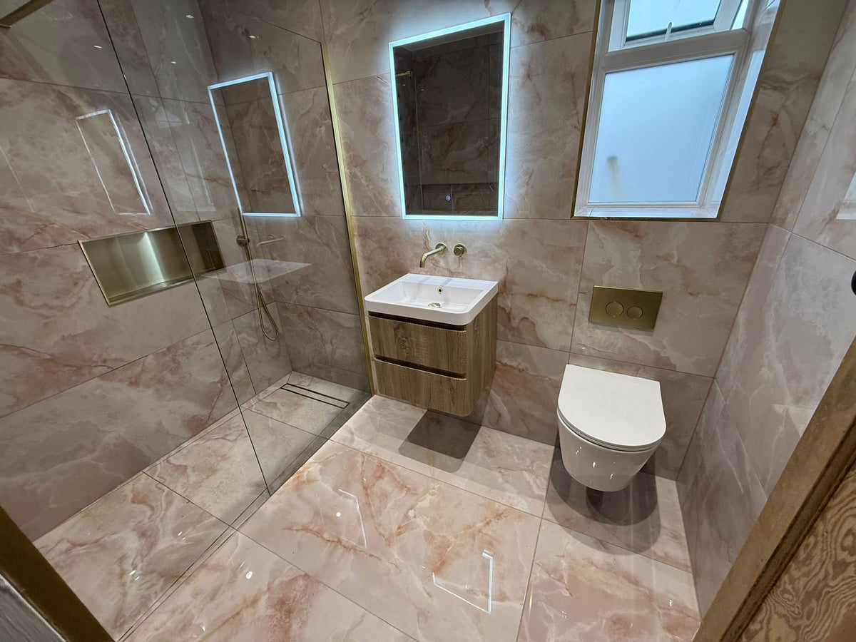 Modern bathroom interior with a glass shower enclosure, wall-mounted sink with illuminated mirror, white toilet, and pink peach marble effect 1200x600 polished  tile on the walls and floor.