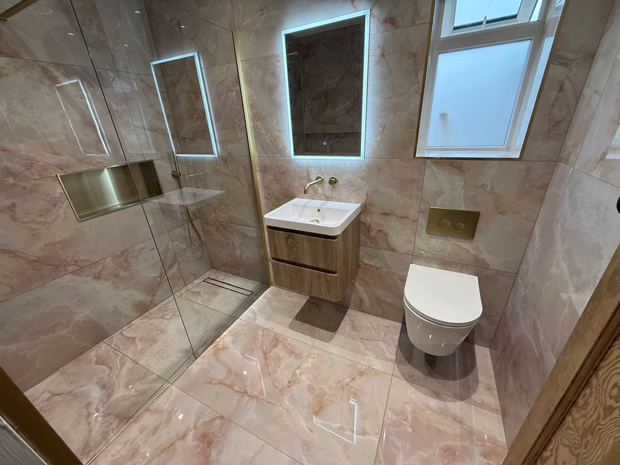 Modern bathroom interior with a glass shower enclosure, wall-mounted sink with illuminated mirror, white toilet, and pink peach marble effect 1200x600 polished  tile on the walls and floor.