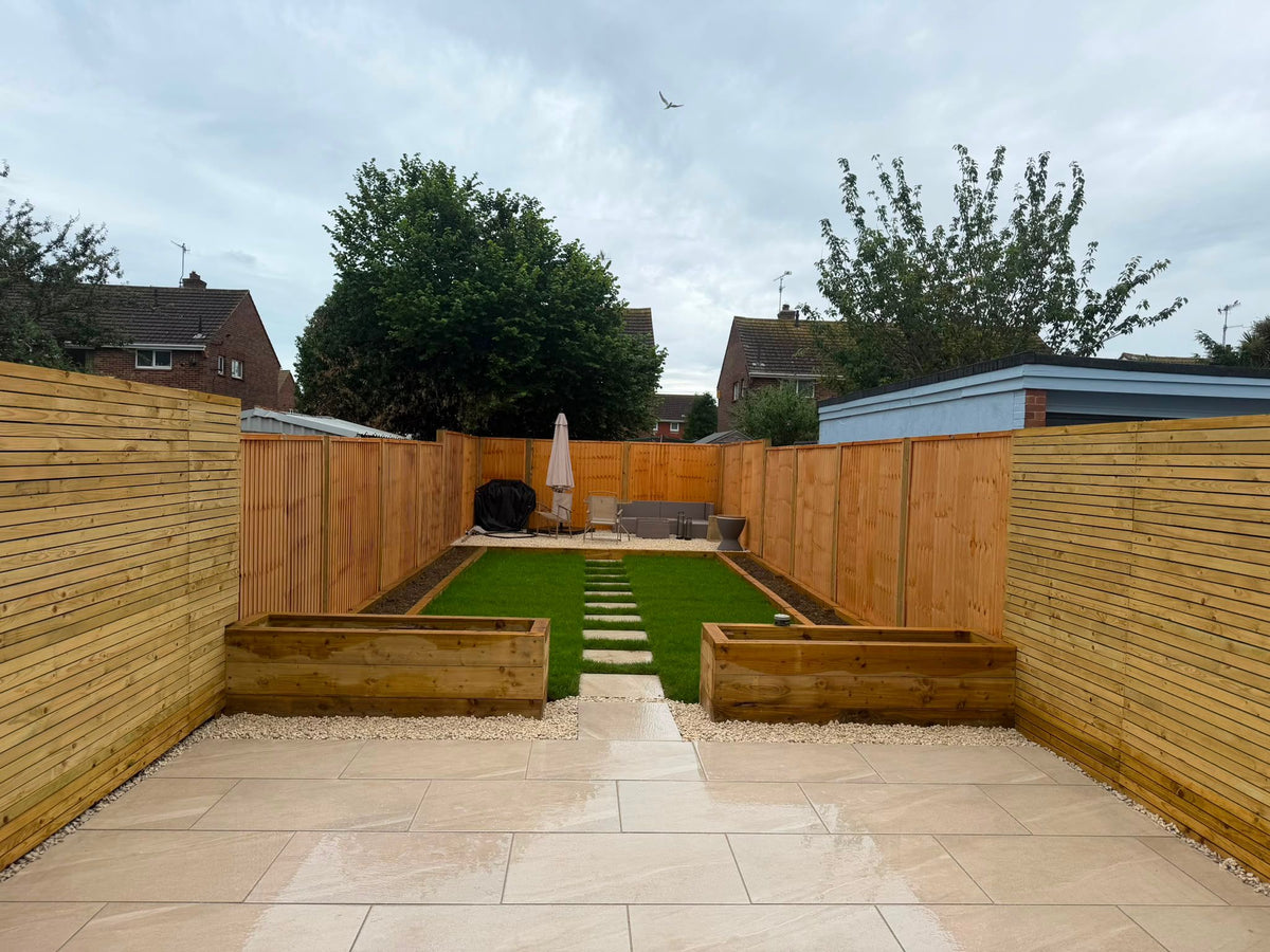 A modern backyard patio and garden setup with light beige stone paving, wooden slat fences, raised wooden planter beds, a green lawn, a stone path, and an outdoor seating area under a cloudy sky.