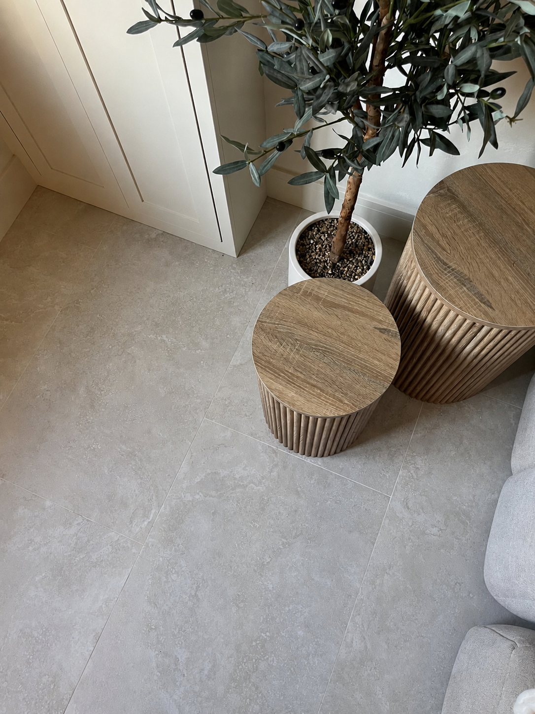 Two round wooden slatted side tables in a modern room setting with a potted olive tree on 1200x600 cream stone effect porcelain tiles