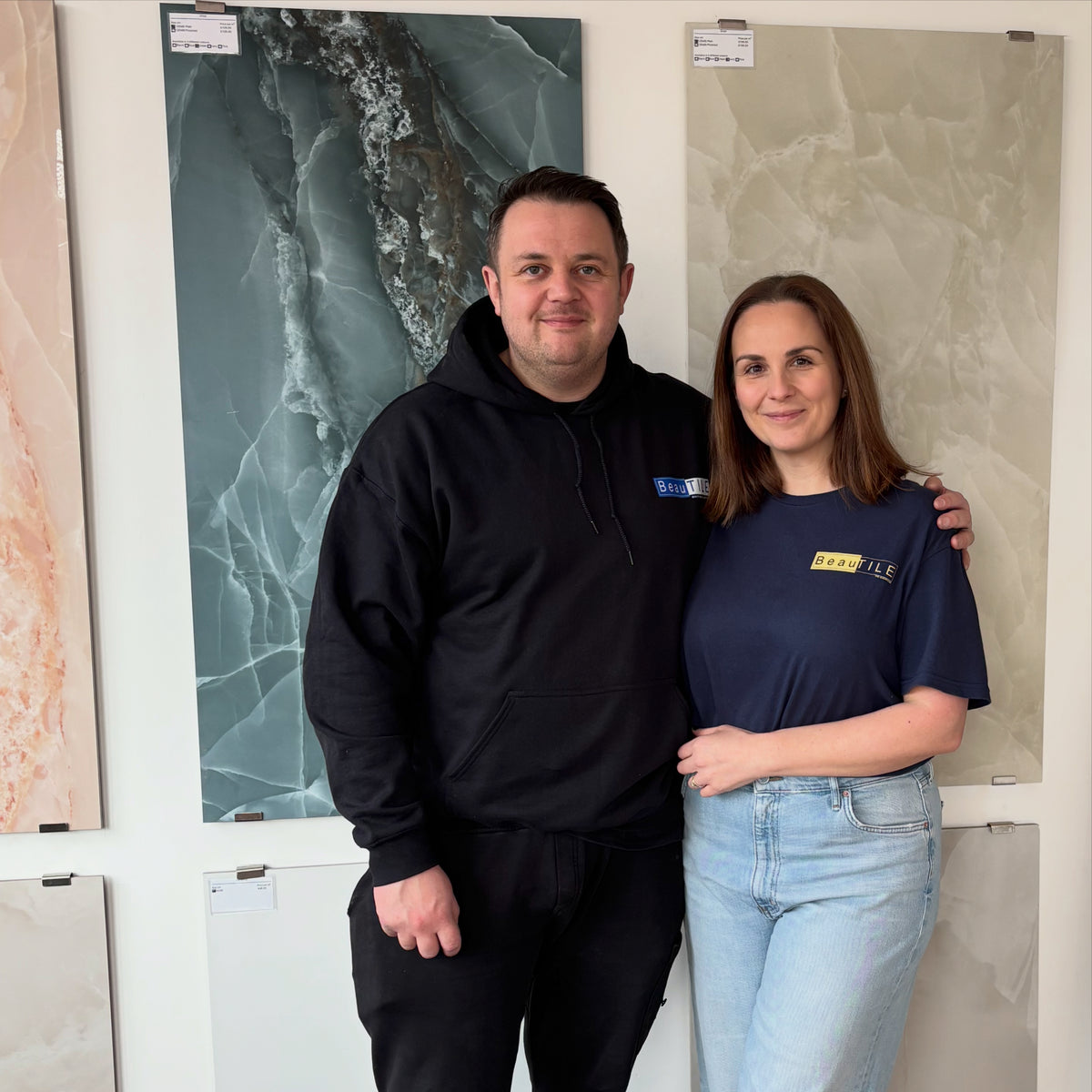 Owners of Beautile, A man and a woman standing together in front of a wall display of large marble or stone tiles. The man wears a black hoodie and black pants, and the woman wears a navy blue t-shirt and light-wash jeans. 