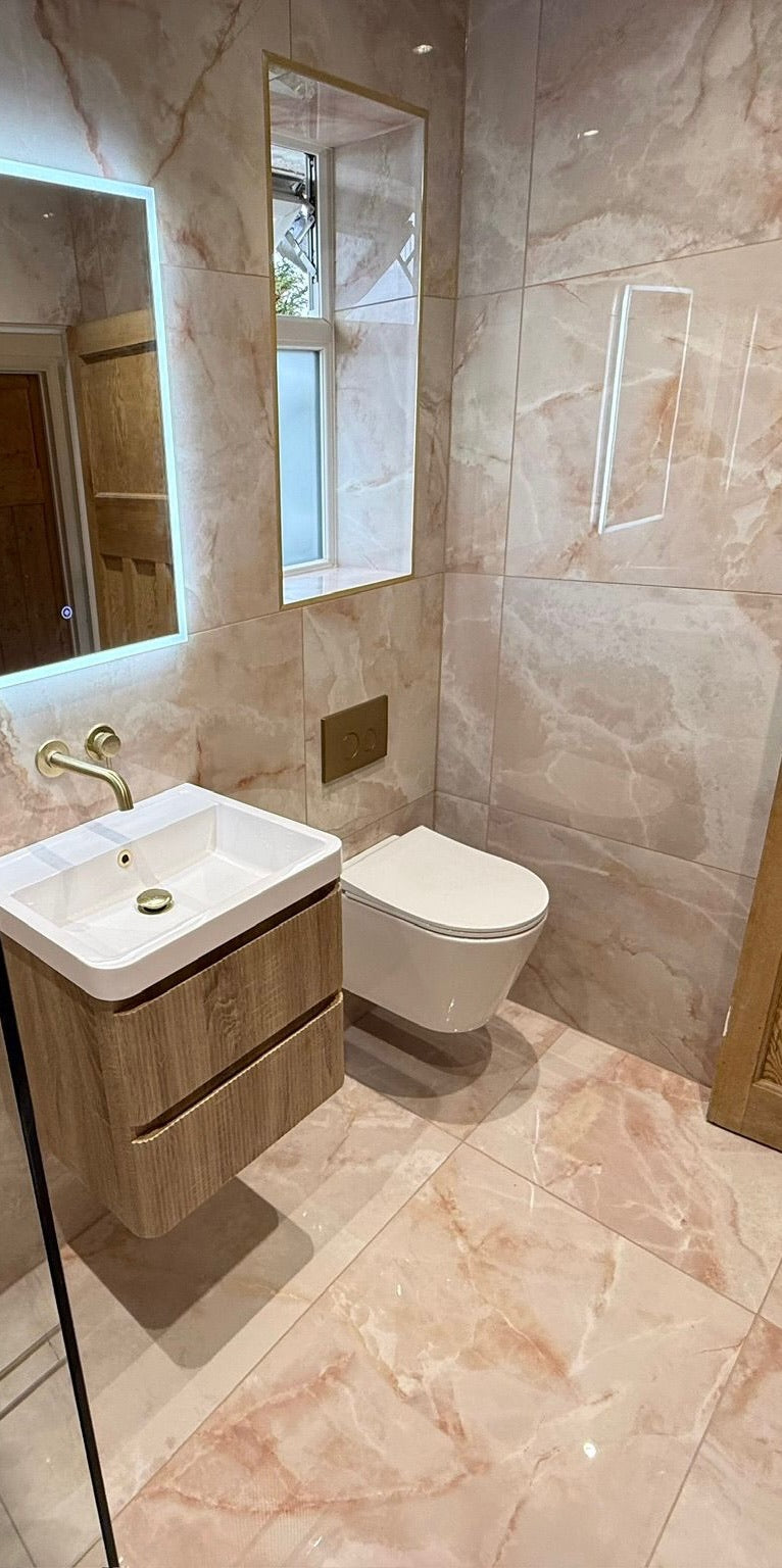 A modern bathroom vanity setup with a white rectangular sink, wood-look floating cabinet, wall-mounted toilet, and a large backlit mirror, all set in pink peach marble-look 1200x600 tiled walls and floor.