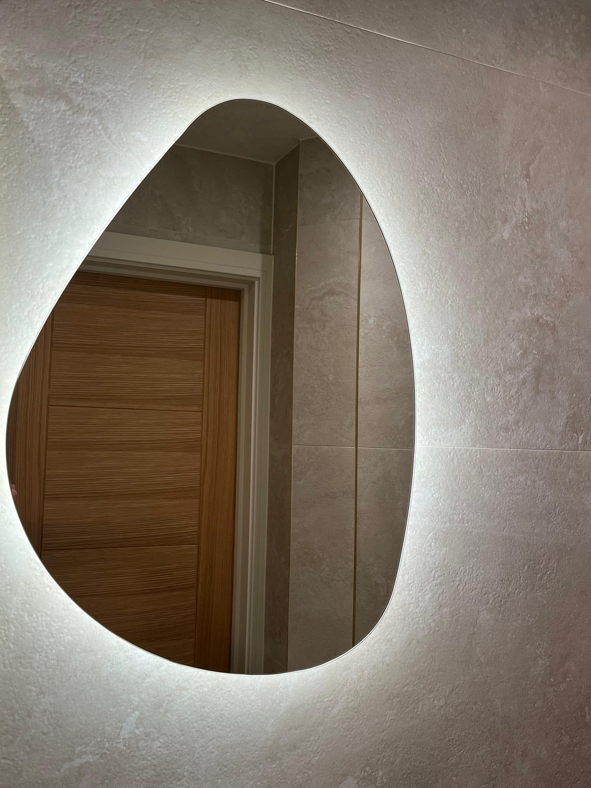 An organic, teardrop-shaped wall mirror with a warm white LED light border, mounted on a textured cream wall and reflecting a wooden door.