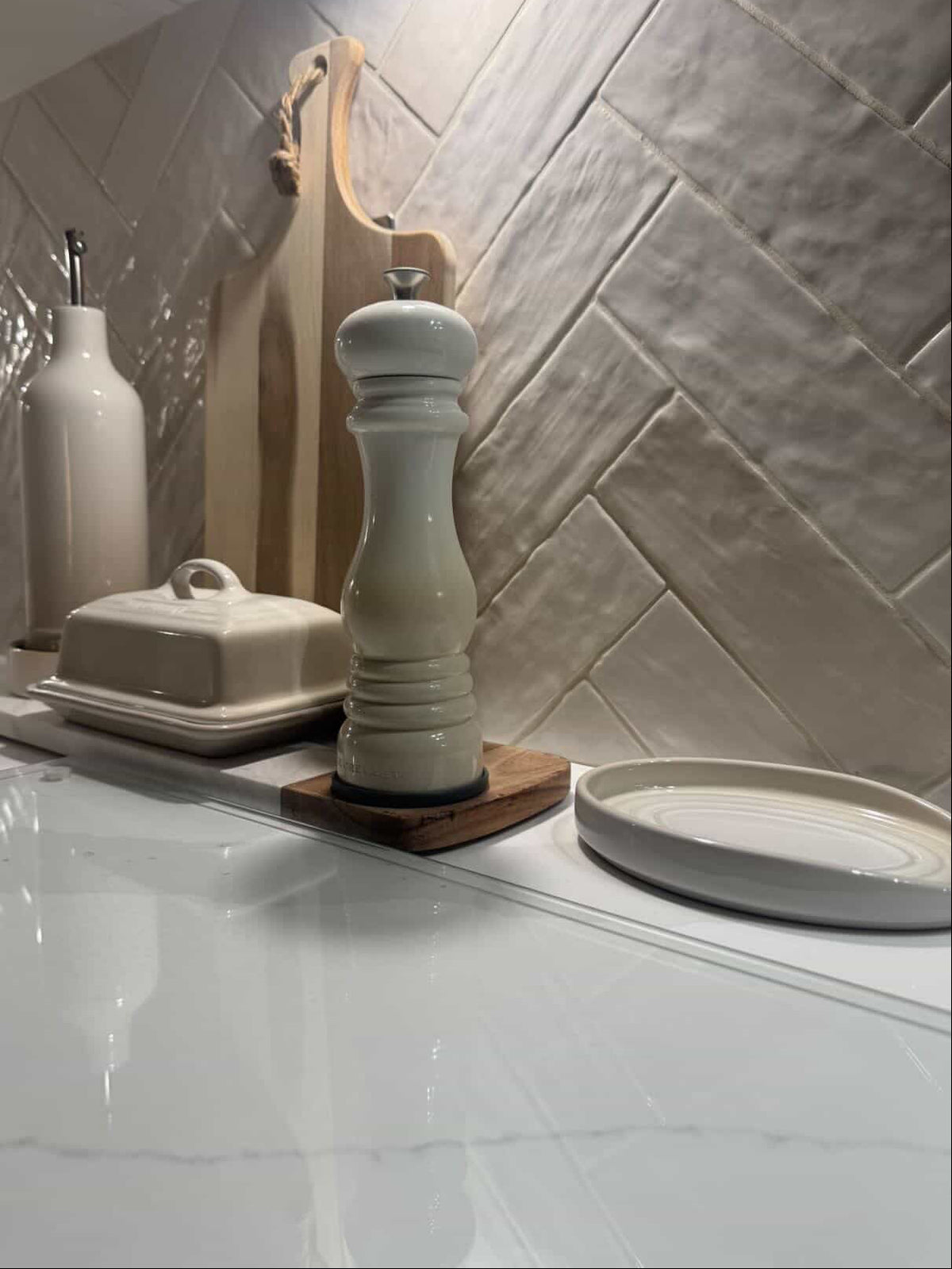 A tall ribbed ceramic pepper mill in a light cream glaze, standing on a small wooden coaster on a white marble countertop. In the background, a textured white herringbone tile backsplash is visible along with other kitchen items including a butter dish, a vase, and a shallow plate.