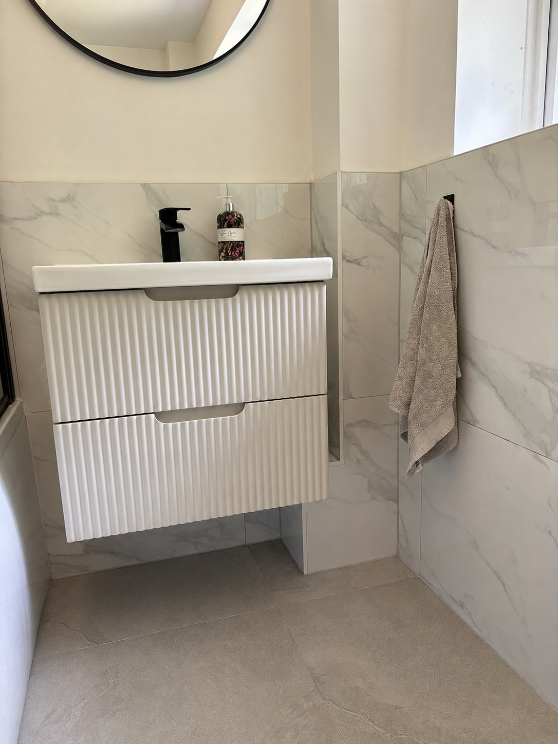 A modern floating bathroom vanity with two ribbed drawers in white, set against marble-look tiled walls and grey tiled floor. A black faucet, soap dispenser, and a hanging beige towel are visible.