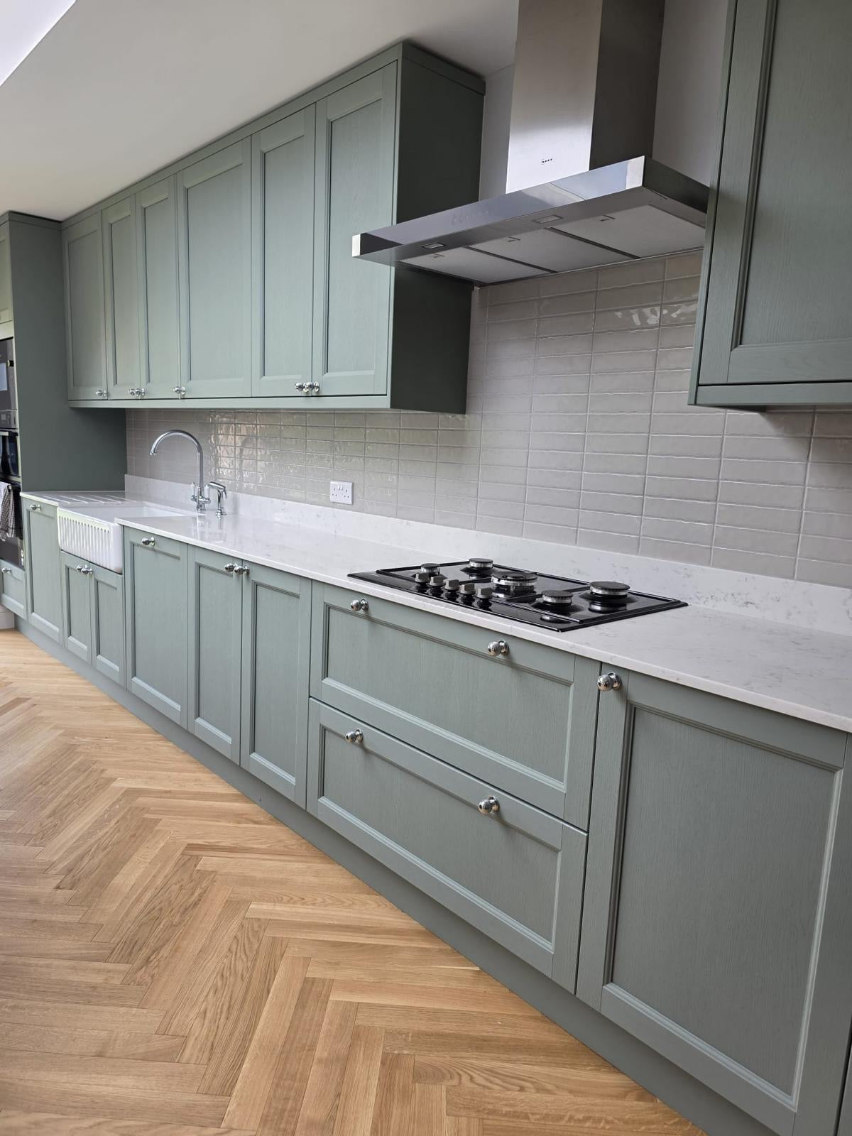 A modern kitchen featuring sage green shaker-style cabinets, a white marble countertop, a black gas hob, a stainless steel range hood, and light wood herringbone flooring.