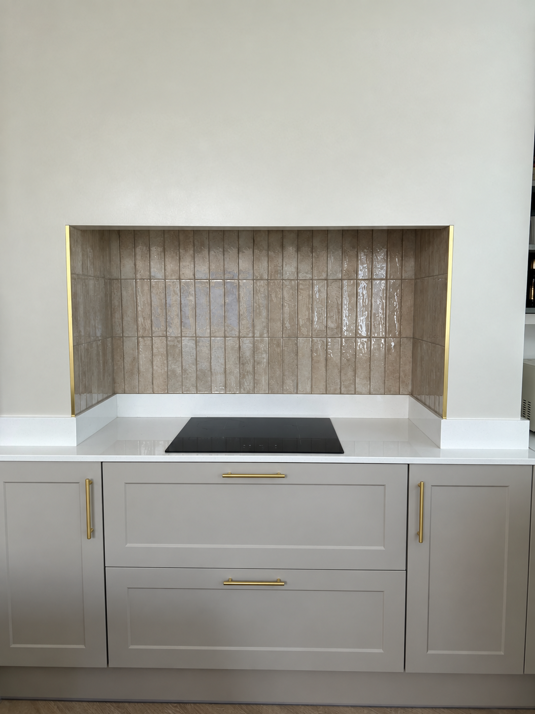 A modern kitchen cabinetry unit with light gray shaker-style cabinets, gold handles, a white countertop, a black built-in cooktop, and a textured beige gloss tile splash back.