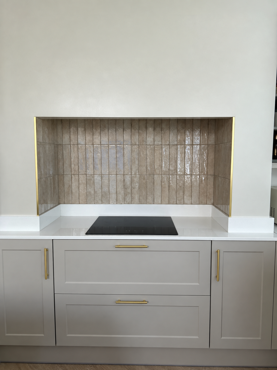 A modern kitchen cabinetry unit with light gray shaker-style cabinets, gold handles, a white countertop, a black built-in cooktop, and a textured beige tile backsplash.