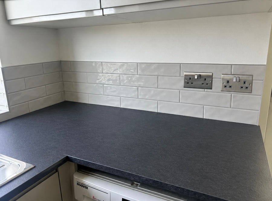 A modern kitchen corner featuring a dark charcoal countertop, a glossy off white subway tile backsplash, and two silver UK wall outlets.