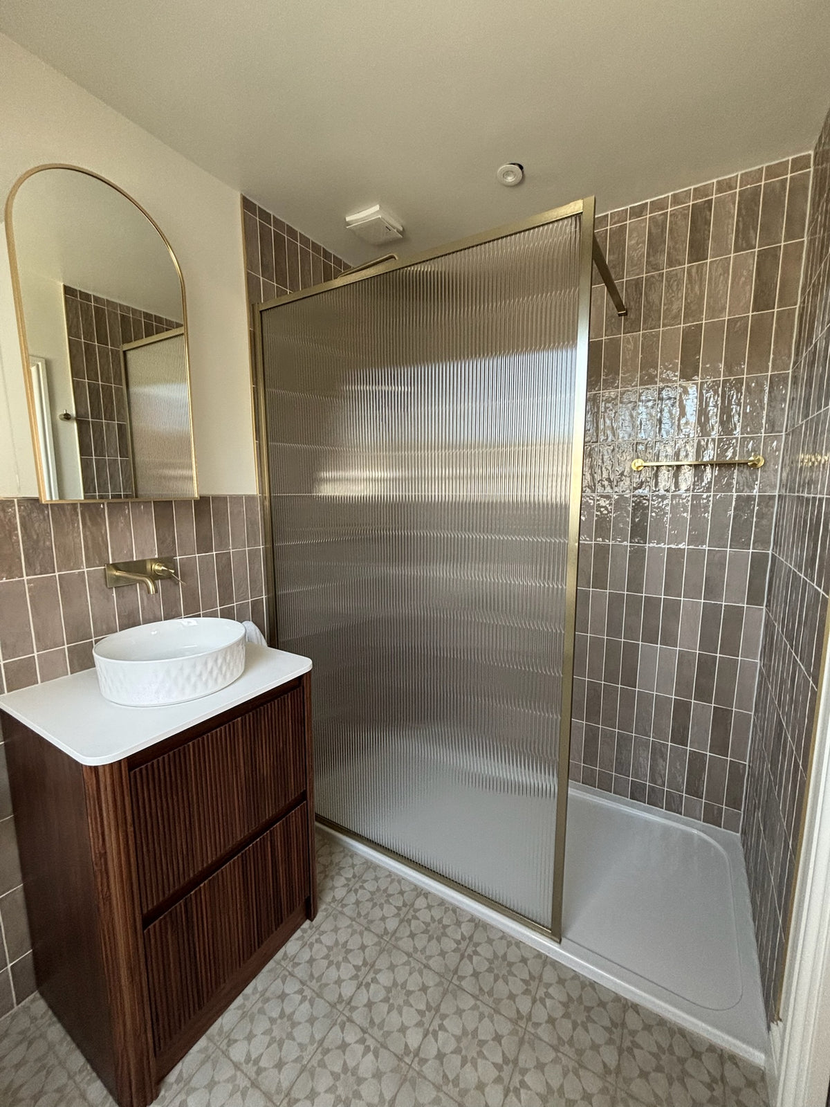 A modern bathroom interior featuring a walk-in shower with a ribbed frosted glass door and gold-toned frame, a dark wood vanity with a white basin, and taupe zeolite style ceramic tiles