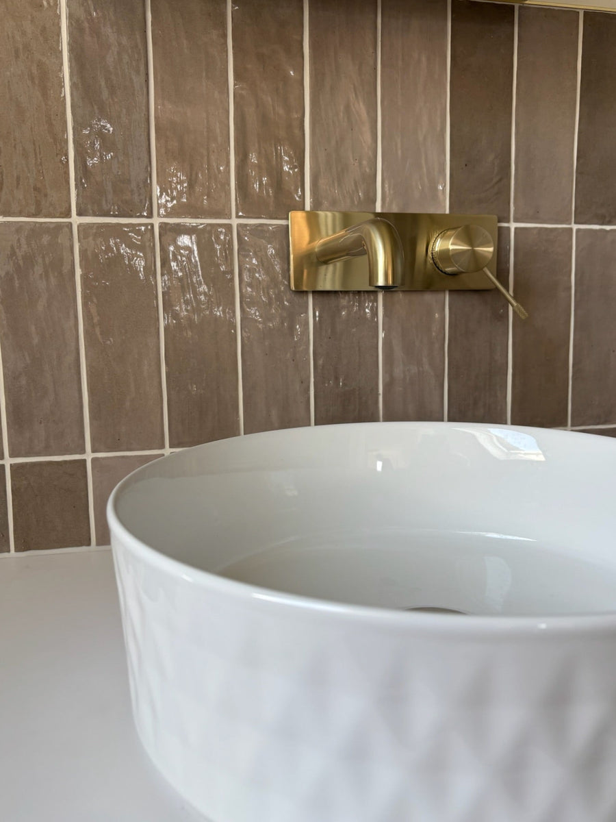 A modern wall-mounted brass faucet with a single lever handle, installed on Taupe glossy, uneven, textured zellige effect tiles with high shade variation above a white vessel sink.