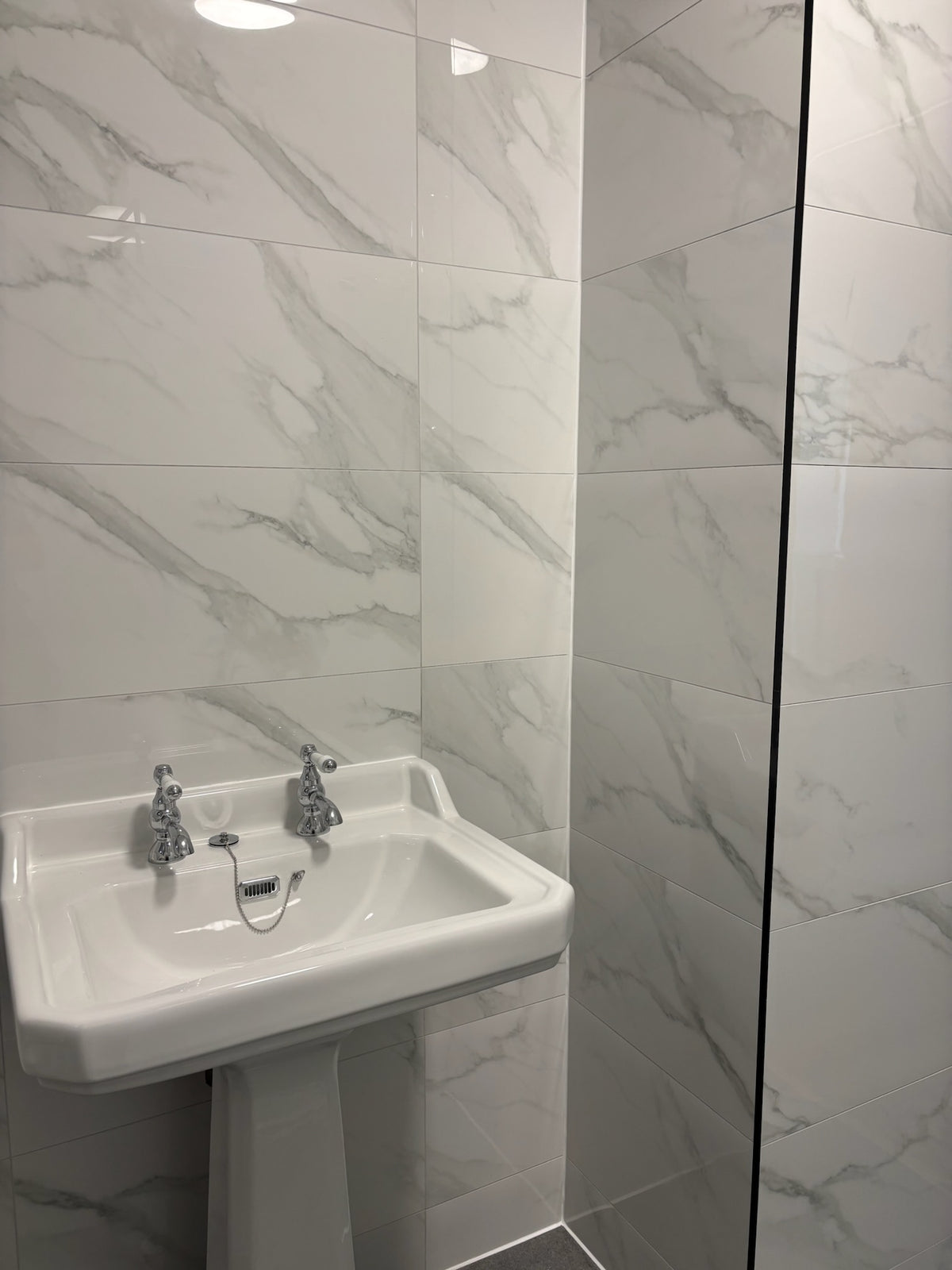 A white ceramic pedestal sink with two chrome faucets installed in a bathroom with white marble-effect gloss wall tiles.