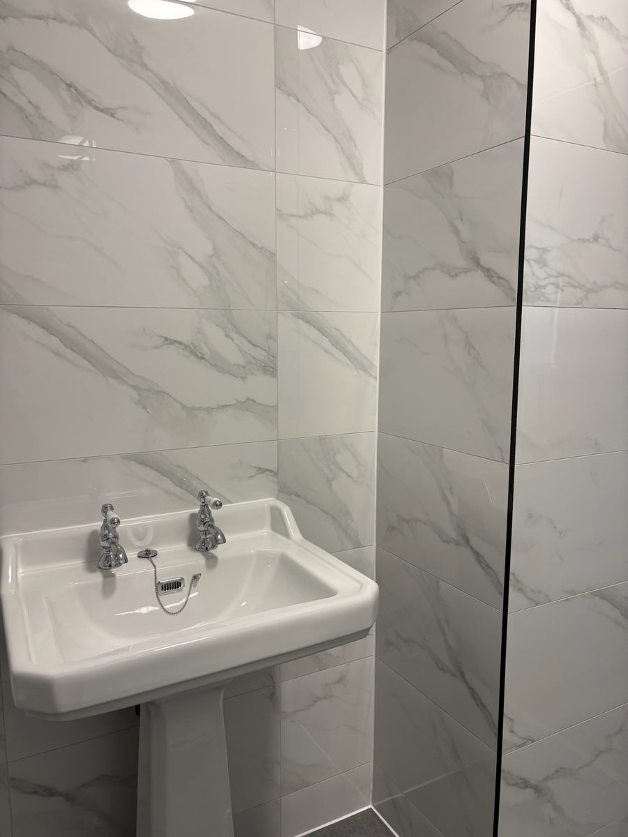 A white ceramic pedestal sink with two chrome faucets installed in a bathroom with white marble-effect gloss wall tiles.