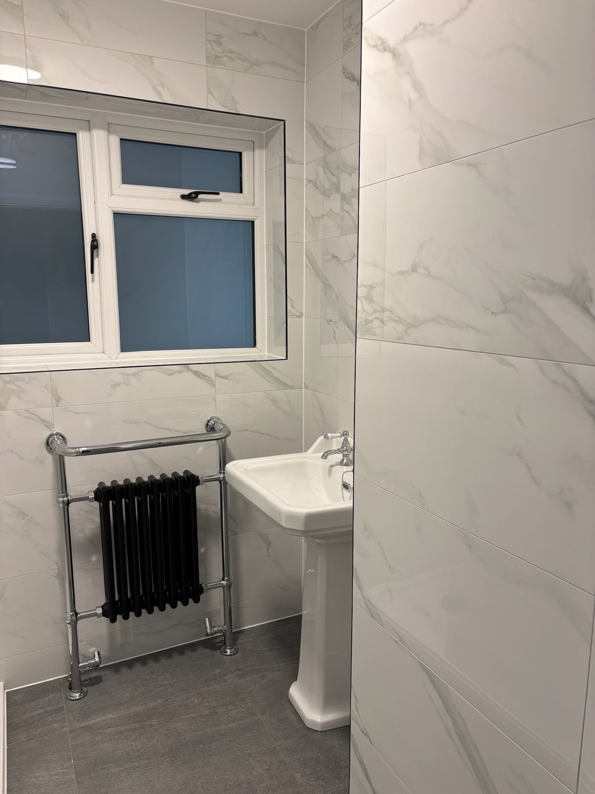 A modern bathroom corner with white marble-effect gloss wall tiles, Anthracite stone effect floor tiles, a white pedestal sink, a window with frosted lower panes, and a chrome towel rail holding a black radiator.