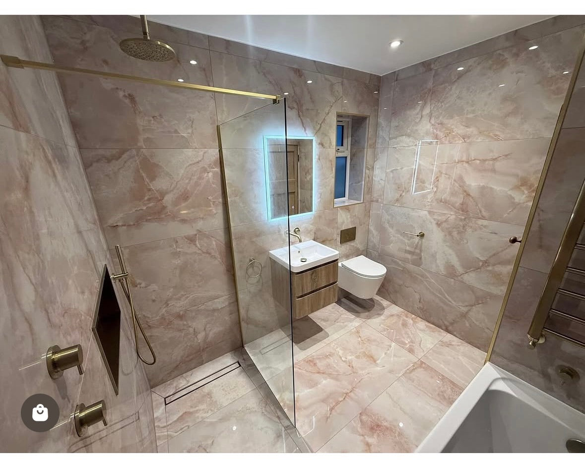 A modern bathroom with pink peach polished marble-look 120x600 tiles, a glass shower enclosure with gold-toned hardware, a white sink with a wood-look cabinet, a white toilet, and a bathtub edge visible in the foreground.