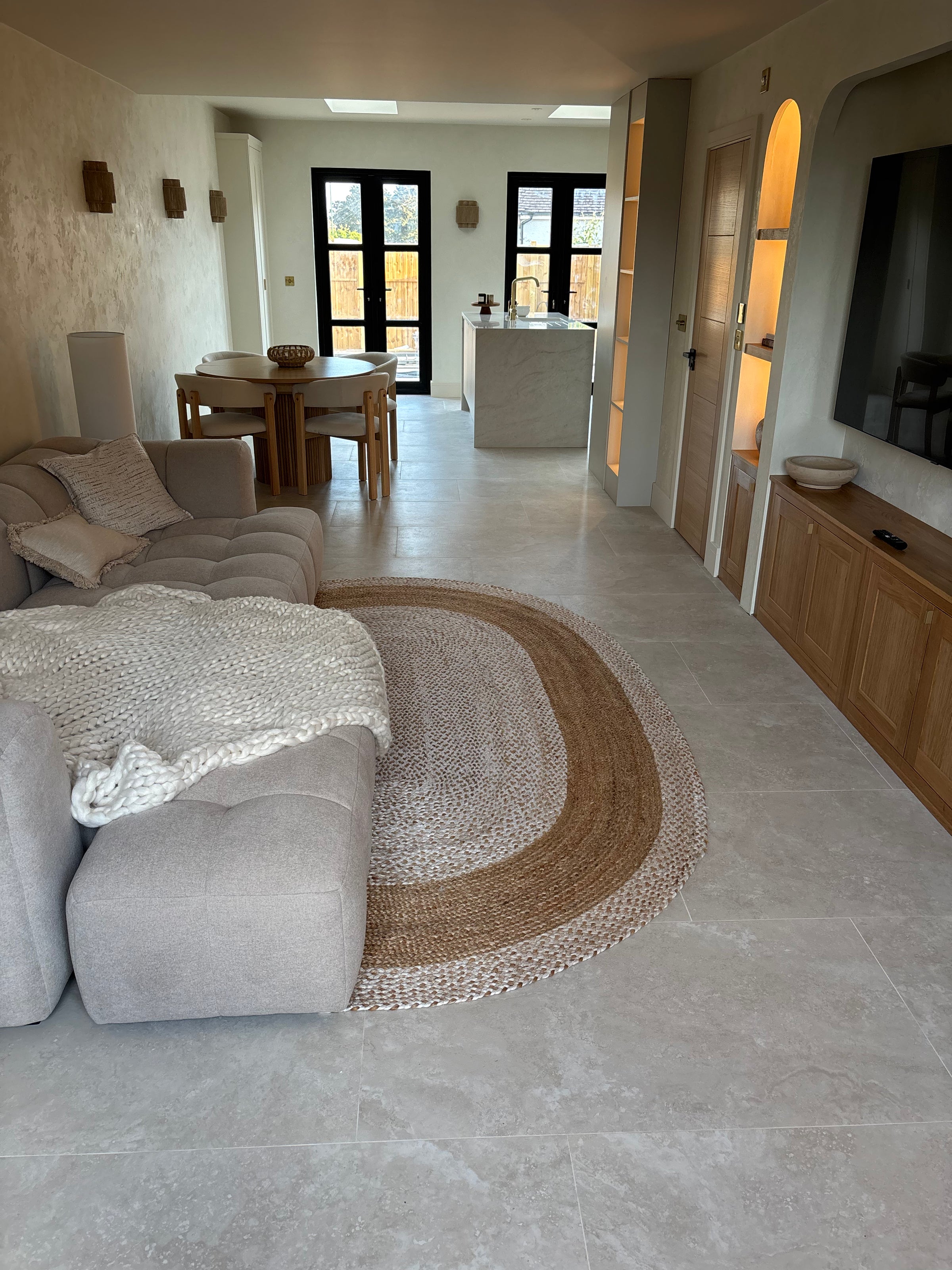 A modern living room featuring a large oval natural jute rug on cream stone effect tile flooring, styled with a light gray tufted sofa and a white knit throw blanket.