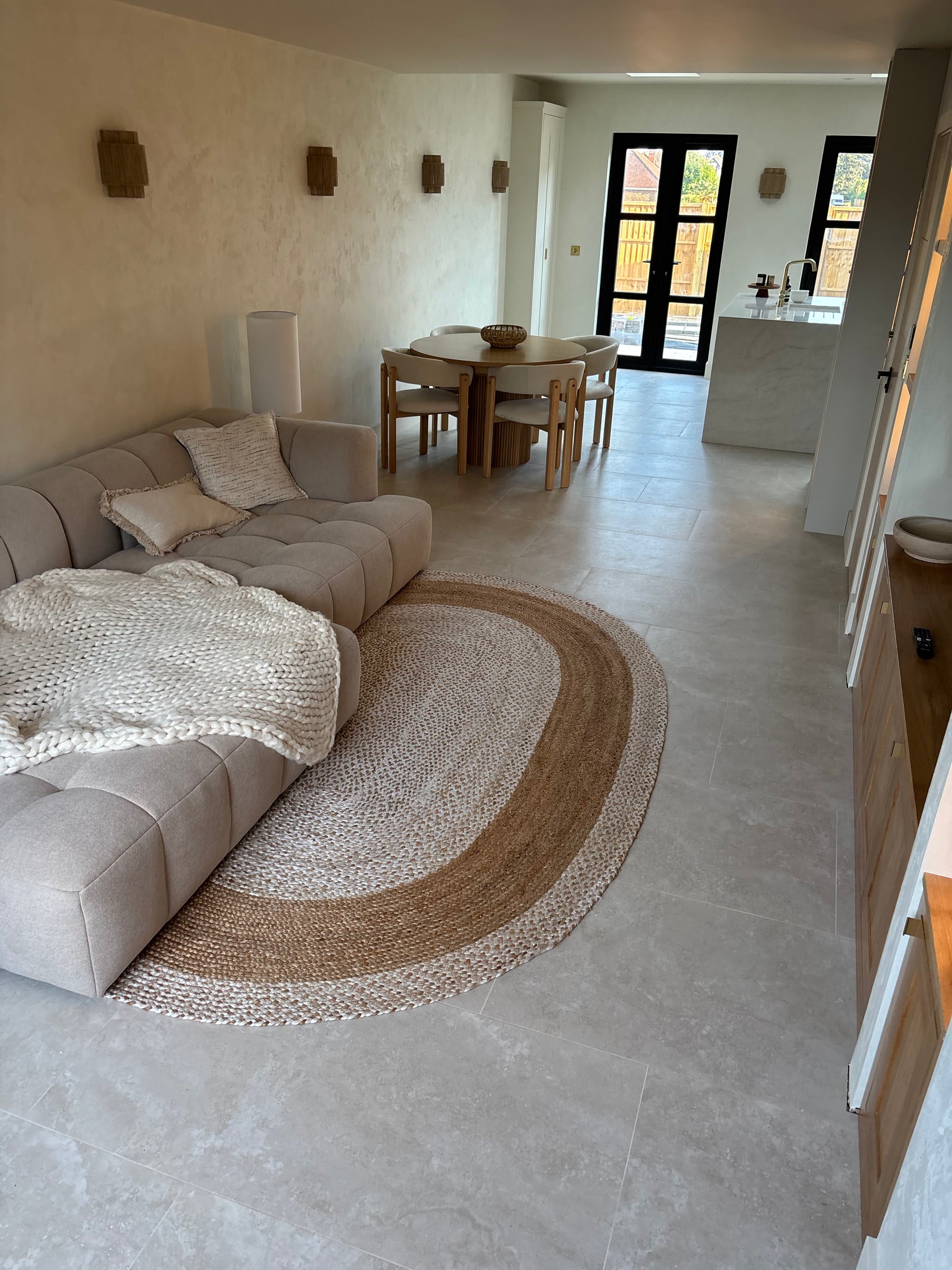 A modern living room featuring a large oval braided jute rug in beige and brown tones, styled under a light gray sectional sofa with a chunky knit cream throw blanket. Tiled in 1200x600 cream stone effect porcelain tiles 