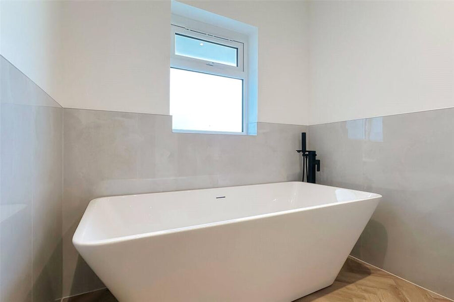 A modern freestanding bathtub in a minimalist bathroom with light grey polished tiled walls, a small window, and a black wall-mounted faucet.