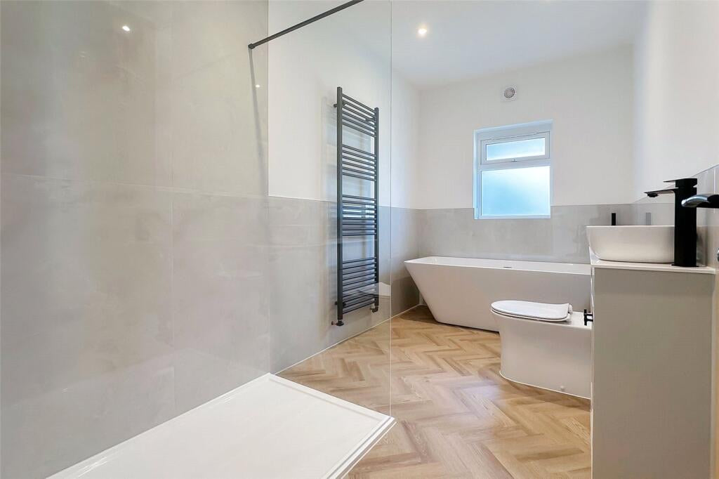 A modern bathroom interior with a freestanding bathtub, white vanity, sink, toilet, wood herringbone floor, grey polished tiled walls, and a glass shower enclosure.