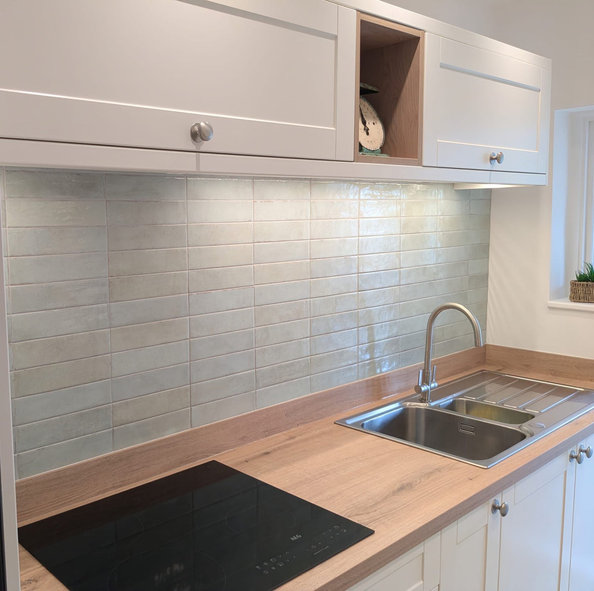 A modern kitchen backsplash featuring glossy, mint green rectangular porcelain tiles mounted on a white wall beneath white shaker-style cabinets and a light wood countertop.